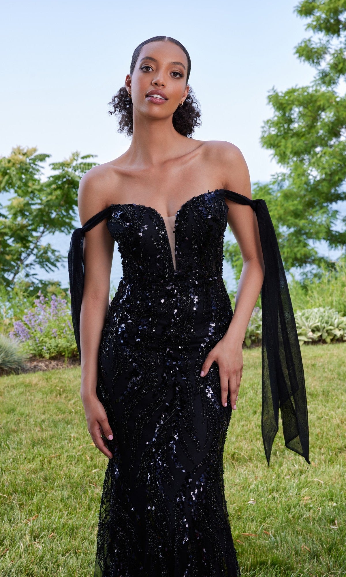 A woman in the Morilee 49166 long prom dress, featuring black off-the-shoulder styling with sparkling embellishments, stands on grass with trees and a tranquil lake in the background.